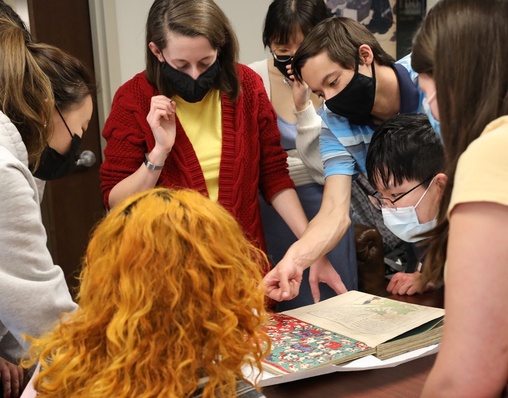 An image of a woman with shoulder length brown hair looking at an open book on a table surrounded by students looking at the book. One male student points to a section of a page.