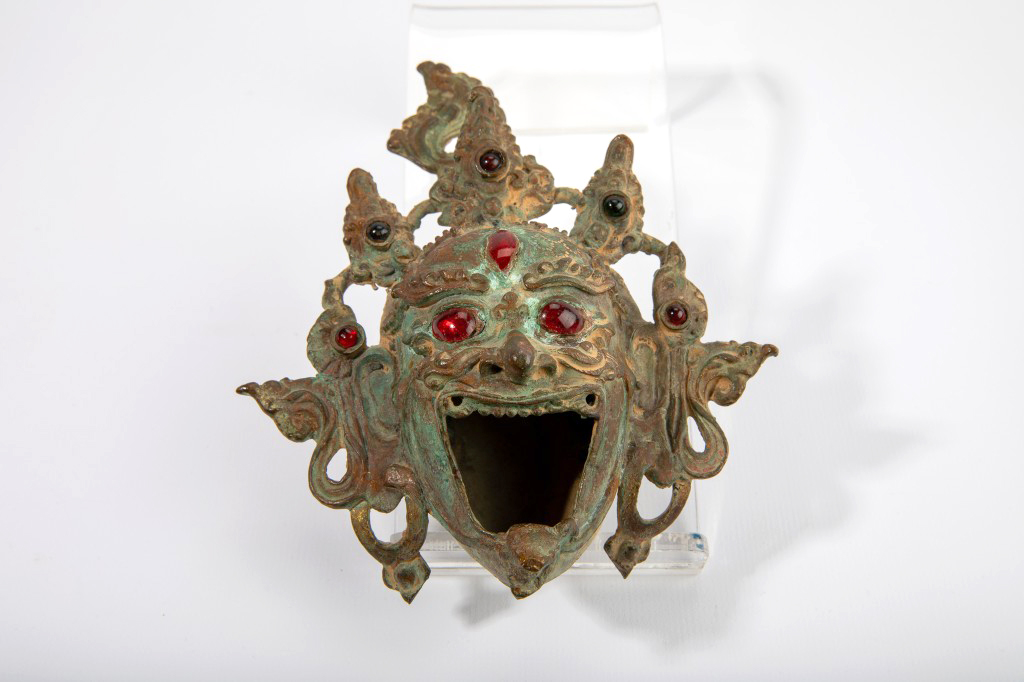 This is a  photo to of a metal incense burner shaped like the head of a Tibetan deity, decorated with red and emerald gemstones.