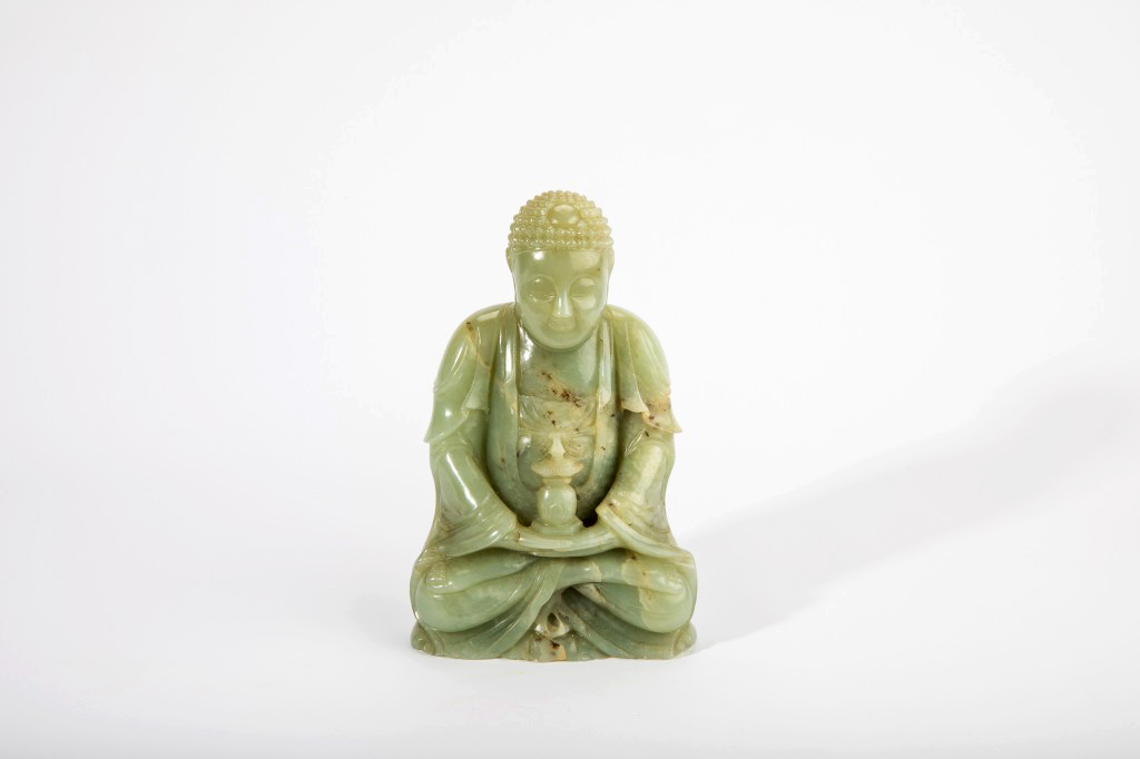 A green jade stone sculpture of the seated buddha with hands holding a stupa form in front of his stomach. The stone has hints of yellow veining throughout.