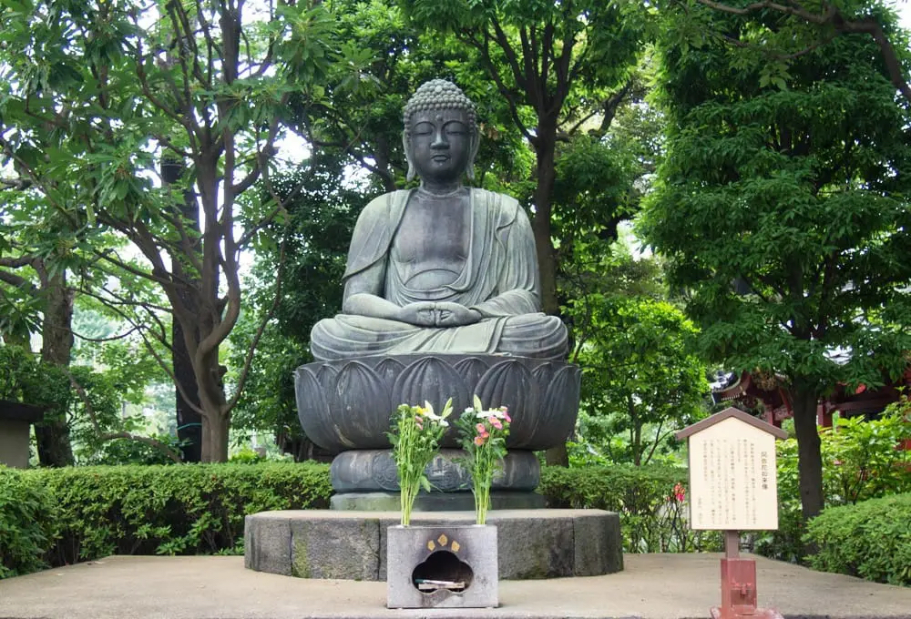 a large seated stone statue elevated on a stone in the middle of the green trees located inside a temple