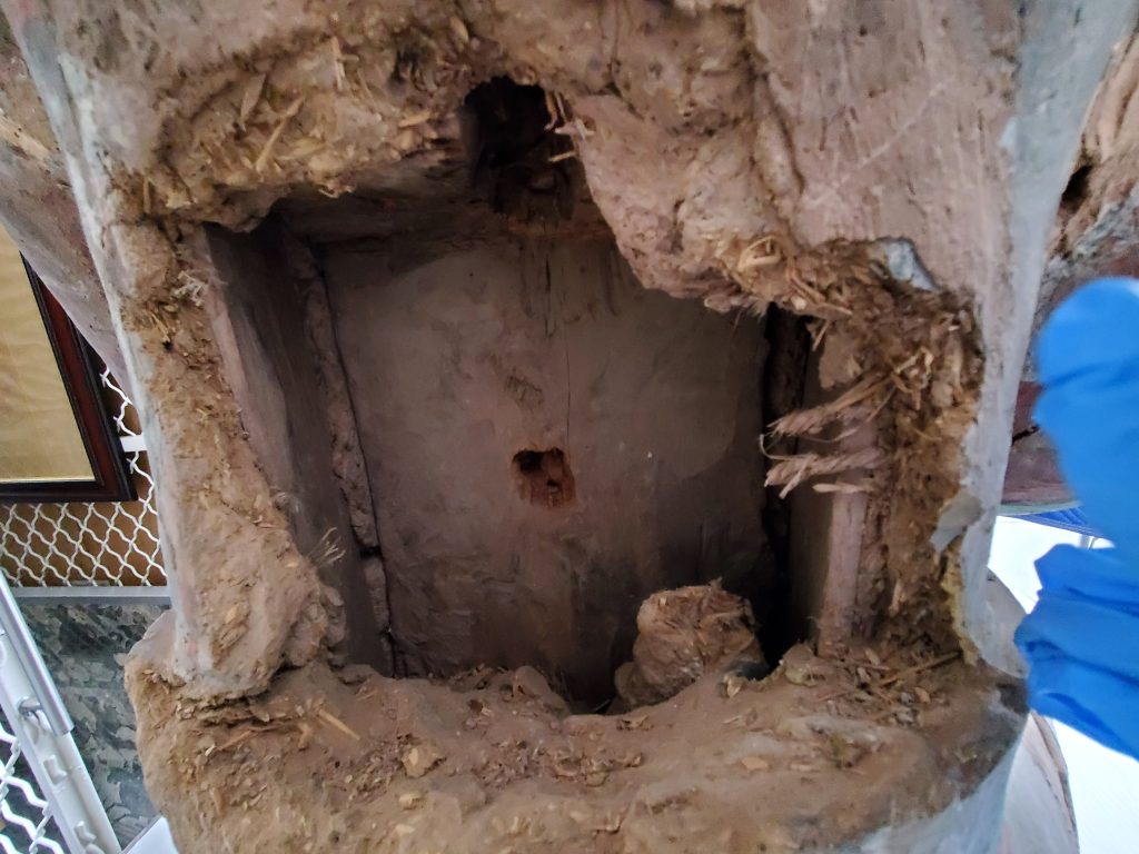 An empty cavity in the back of the seated Bodhisattva statue. The opening is irregularly shaped with straw and other organic materials sticking out of the terracotta. There is a wooden panel at the back of the cavity with a circular indentation in its center.