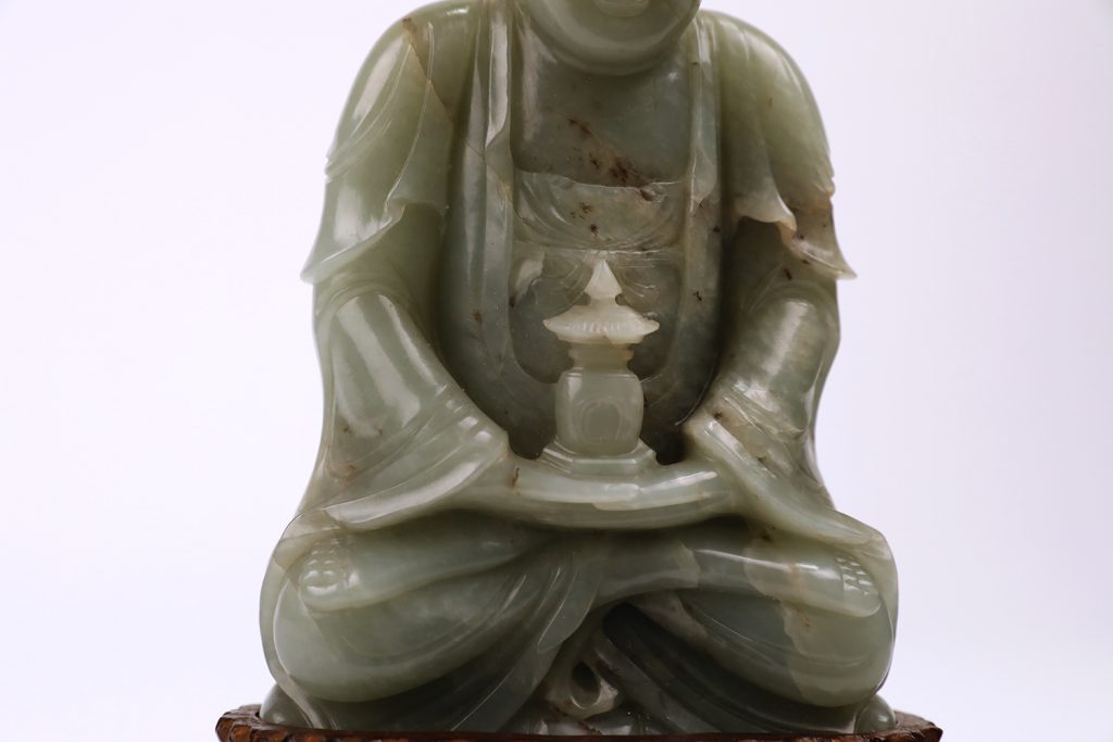 detail view of a green stone seated buddha figure showing hands on top of folded legs holding a small vessel in the shape of an architectural form like a stupa 