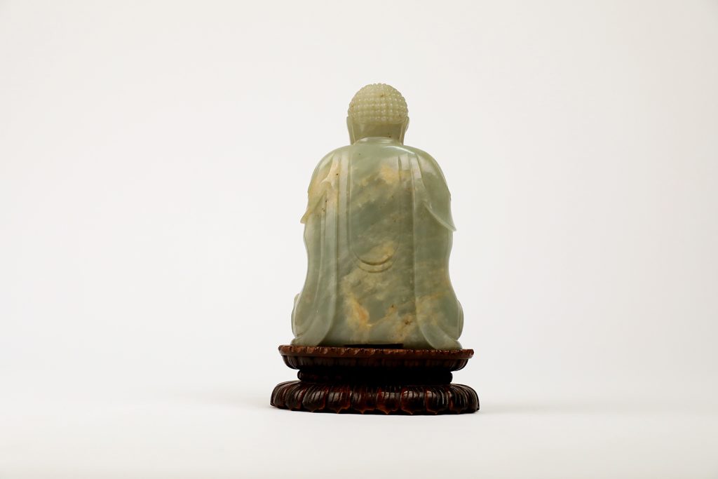 View of the back of a jade seated buddha figure on a small wooden platform. The jade is green with yellow veining.