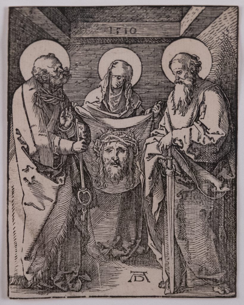 Two men stand on either side of a woman who holds a large cloth with the image of Jesus’ face imprinted on it.